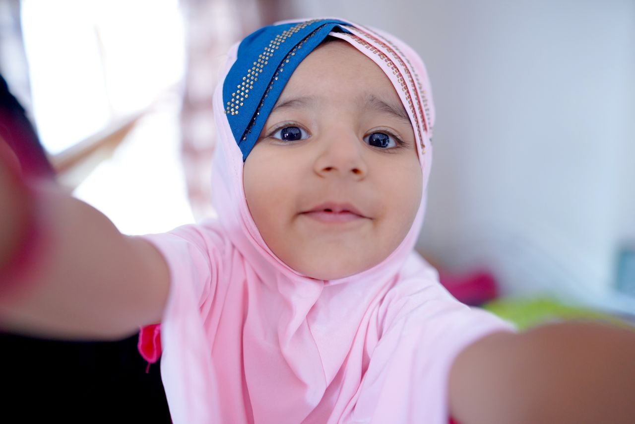 Adorable child wearing a pink hijab, smiling while taking a selfie indoors.